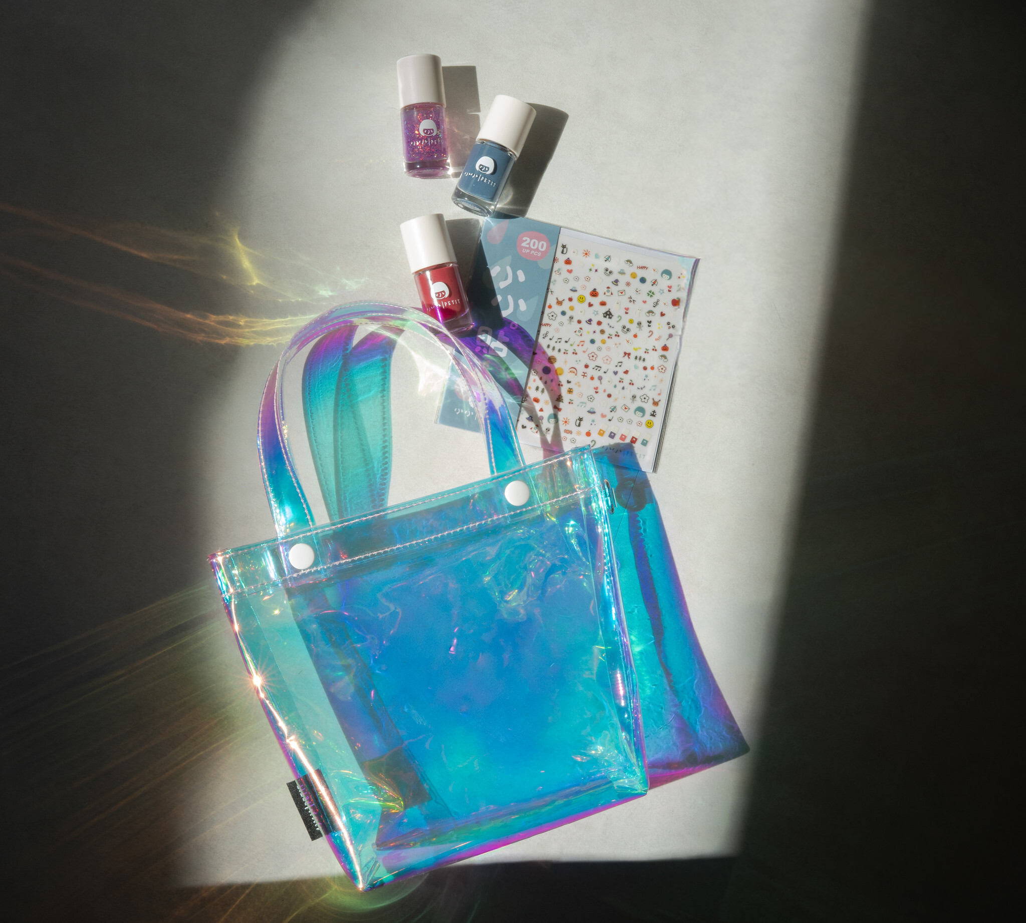 小小PETIT × 理理 liil 聯名小小微光包｜透明虹光托特包搭配三款無毒水性可撕指甲油與小小貼貼，置於自然光影下的聯名限定組合平拍圖