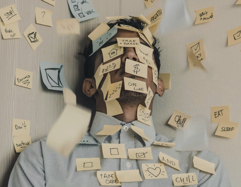 一位穿著襯衫的男子被黃色和藍色的便利貼完全覆蓋臉部和上半身,便利貼上寫著「STOP」、「TAKE A BREAK」、「CHILL」等提示,象徵著工作過度、壓力大或待辦事項淹沒