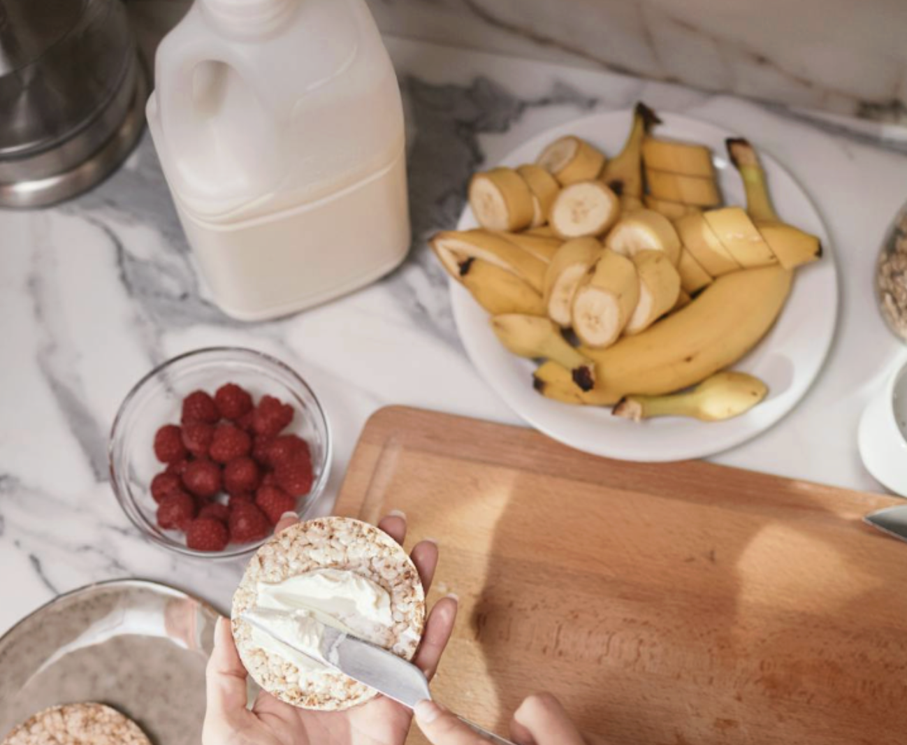 從上方俯視，呈現準備健康早餐的畫面，包含一盤切片的香蕉、一碗紅莓，以及手持奶油刀在米餅上塗抹奶油乳酪，旁邊擺放著一罐牛奶