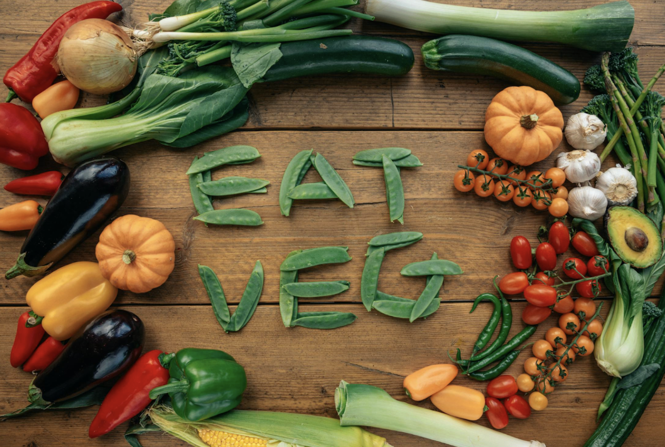 俯視拍攝的木質餐桌上，擺滿了茄子、甜椒、玉米、南瓜、番茄、酪梨等多種新鮮繽紛的蔬菜，中間用豌豆莢排出「EAT VEG」的文字，提倡健康素食或多吃蔬菜的飲食習慣