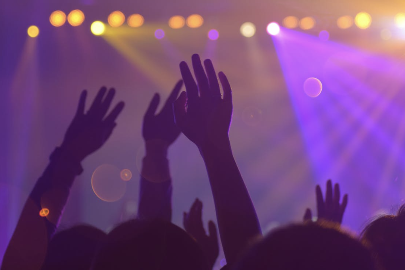 演唱會或音樂節現場,觀眾將雙手高舉向舞台,背景充滿紫色、黃色光束和聚光燈效果,營造出熱烈、興奮的派對或表演氣氛