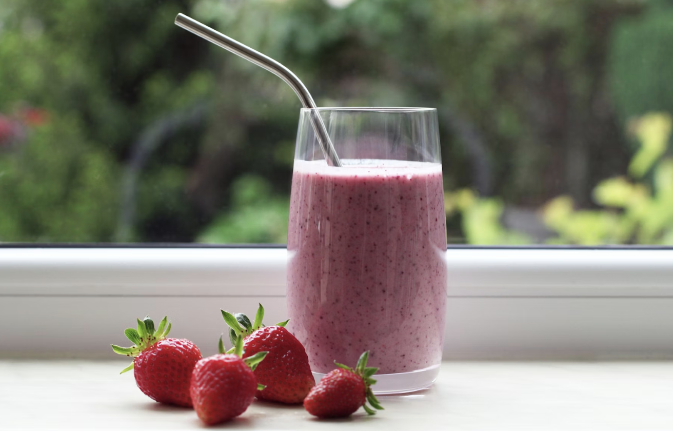 一杯粉紫色草莓果昔放在窗邊桌面,搭配不鏽鋼吸管與新鮮草莓,呈現清新自然的健康感