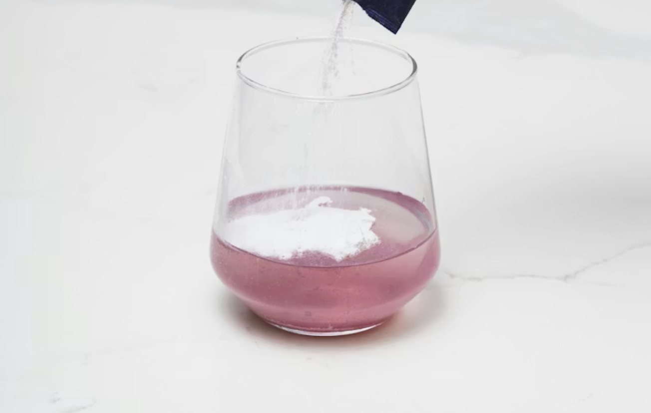 透明玻璃杯中盛裝淡紫色飲品,白色粉末從上方倒入液體中,呈現飲品沖泡過程的瞬間