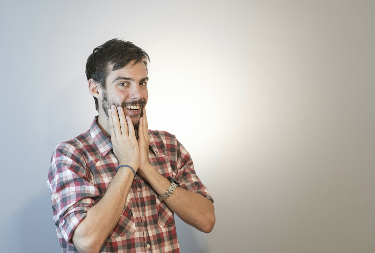 一名留著鬍鬚的年輕男子雙手輕觸臉頰微笑,表情驚喜又愉悅,背景簡潔明亮