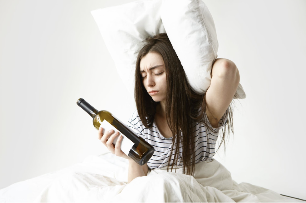 一位長髮女子神情痛苦地坐在白色床鋪上，她一手拿著空酒瓶，另一手用枕頭壓著頭部，展現出宿醉或派對後嚴重頭痛的不適感。