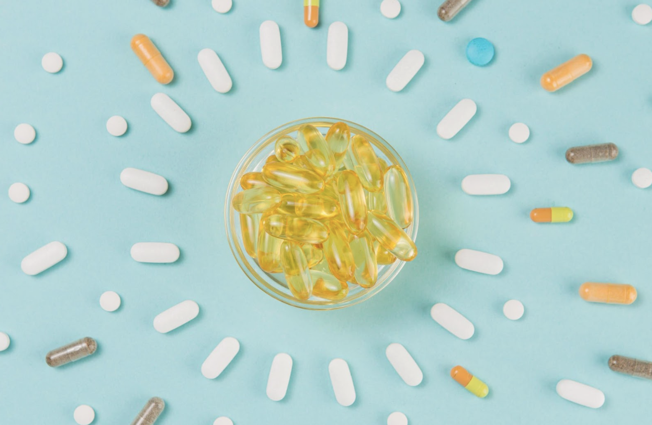俯瞰視角呈現淺藍色背景,中心是一個裝滿金黃色魚油膠囊的小玻璃碗,周圍由各種白色藥錠、彩色膠囊(橘、綠、灰等)以放射狀整齊排列,呈現出醫藥、營養補充或健康管理的意象。