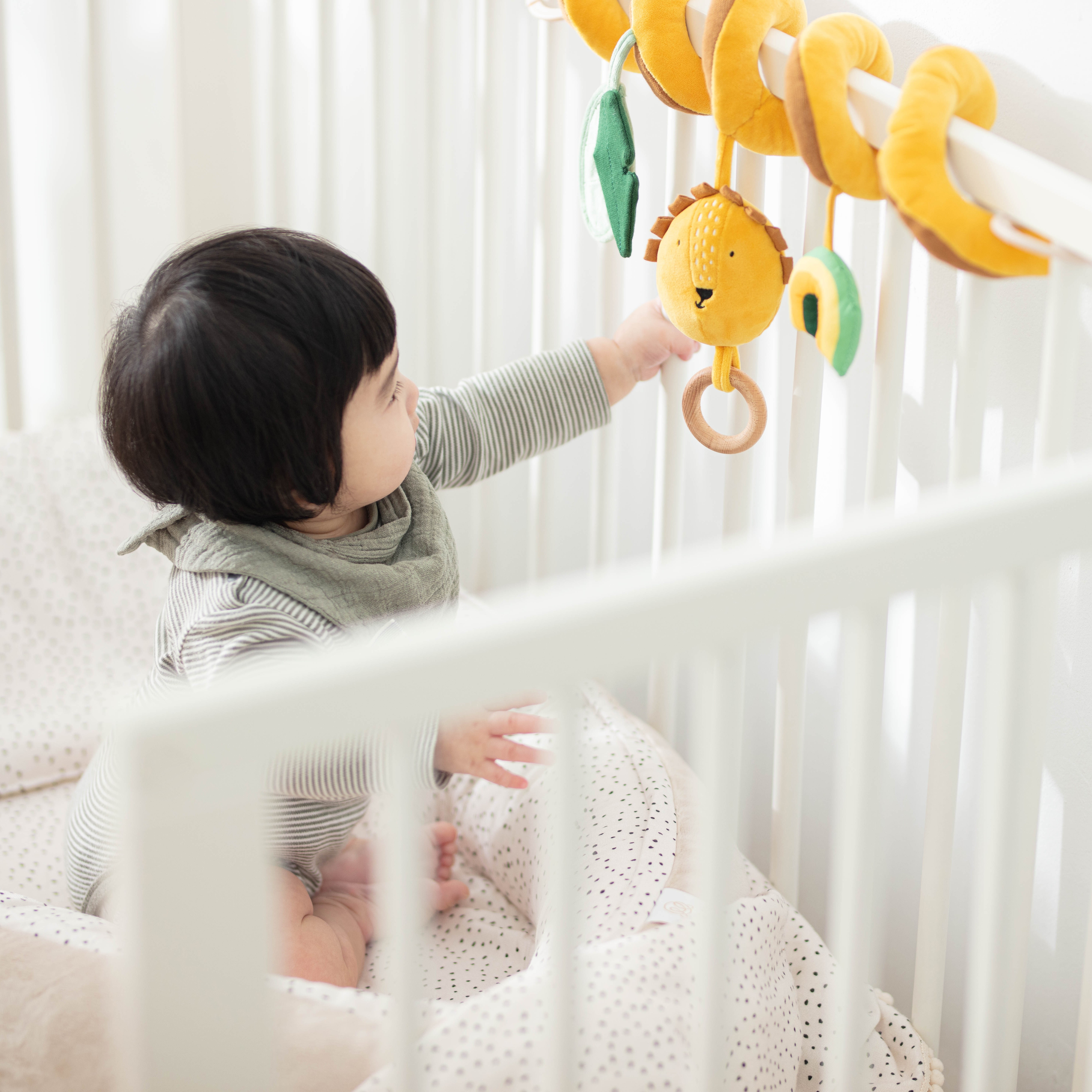 寶寶在嬰兒床內抓取圍欄上的比利時 Trixie 陽光獅子造型捲捲安撫玩具，布質材料安全防護。