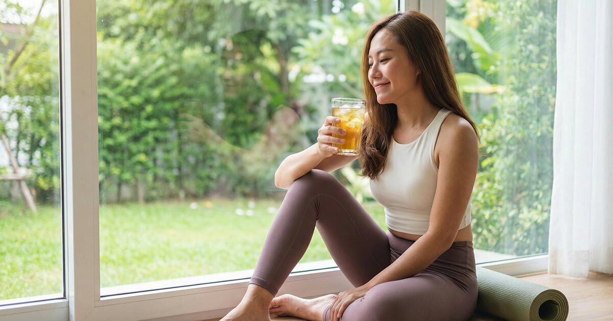 一位女性在窗邊瑜珈運動後，放鬆地享用一杯加冰塊的冷萃黃金蕎麥茶，體現無咖啡因的健康生活與身心平衡。