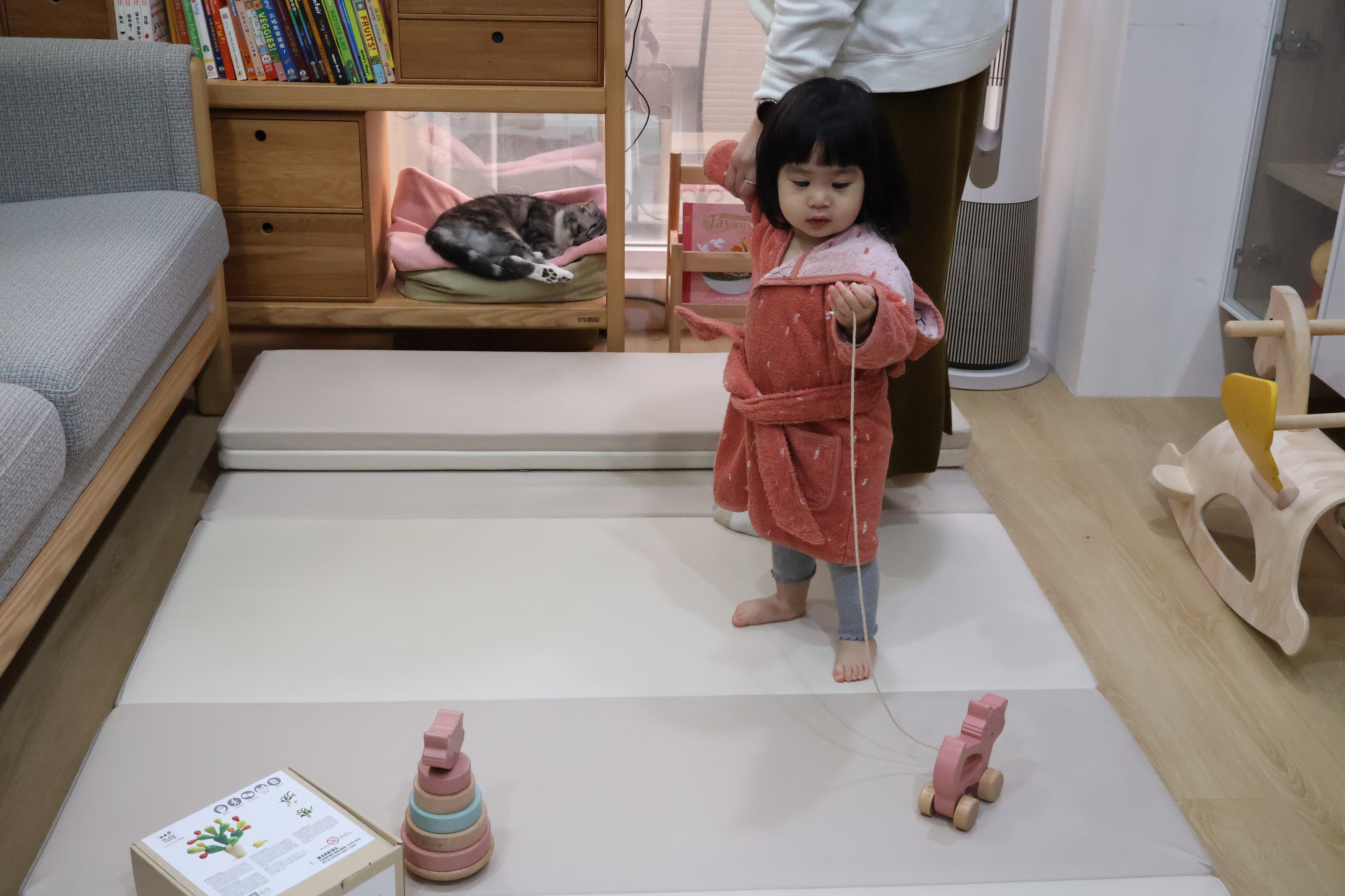 小梅子穿著粉色浴袍，在客廳地墊上拉著貓咪造型的寶寶木玩拉車。