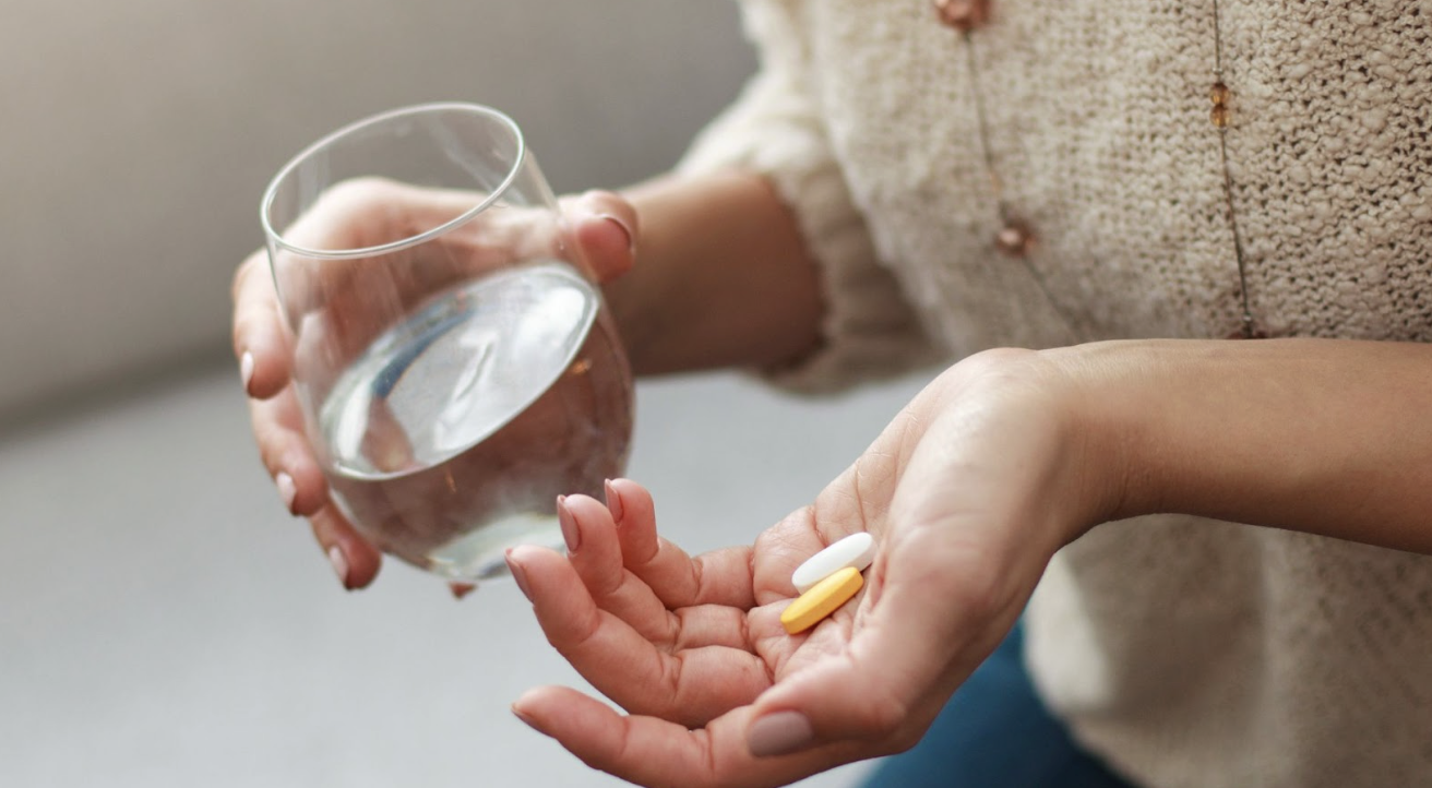 一名女子一手拿著裝水的玻璃杯，另一手掌心放著兩顆藥錠（一白一黃），準備服藥的特寫畫面