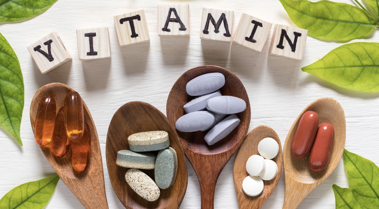 五支木湯匙上盛放著各式形狀與顏色的營養補充品（包括膠囊、錠劑與軟膠囊），上方排列著拼出「VITAMIN」字樣的木製方塊，背景點綴著綠色葉片，呈現健康與維生素補充的概念。