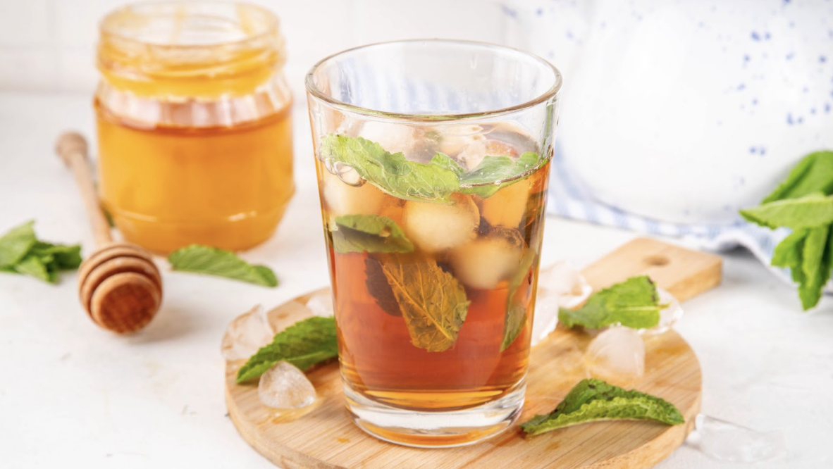 一杯裝有冰塊與新鮮薄荷葉的冰茶,背景擺放著一罐金黃色蜂蜜及木製蜂蜜攪拌棒,呈現清爽、天然調味的飲品意象