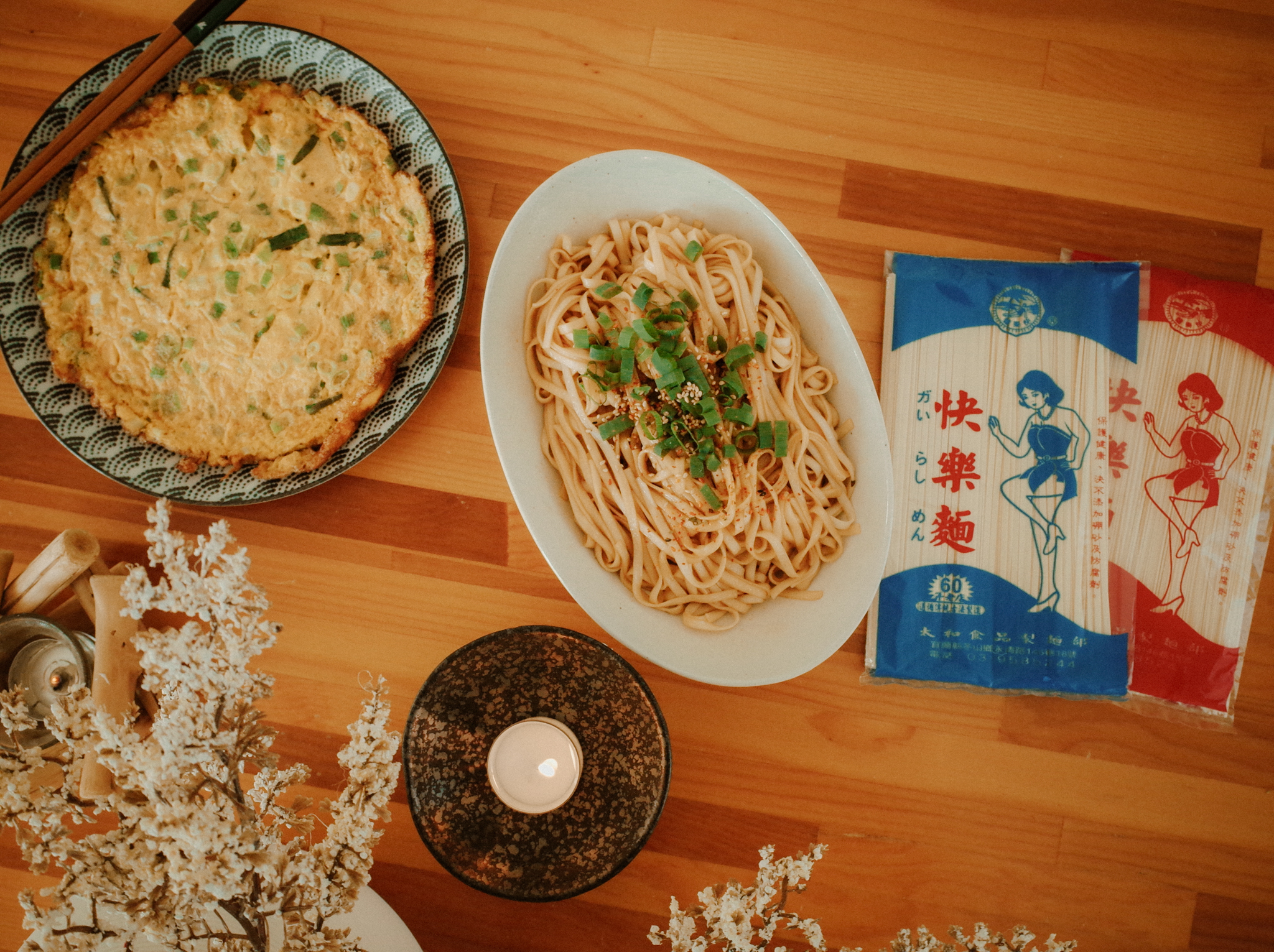 日式風格木桌上的美味料理，包含蔥蛋、三星蔥油拌麵、復古包裝麵條與點燃的香氛蠟燭。