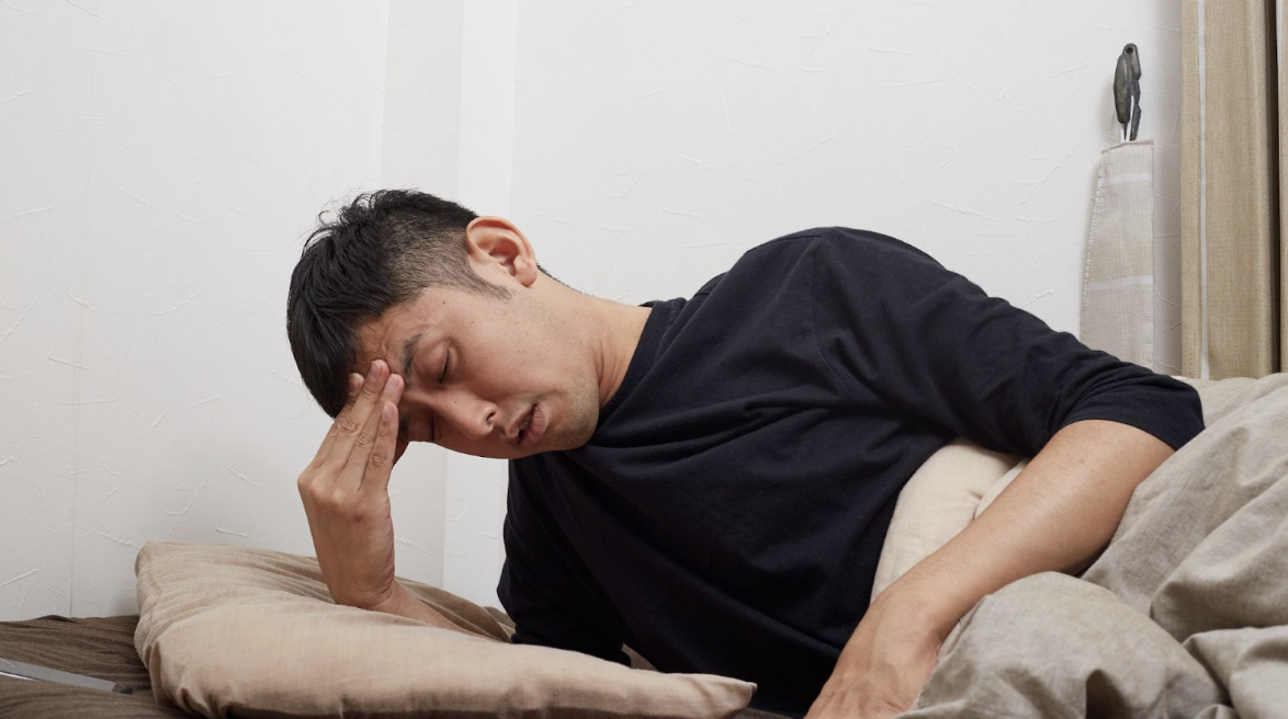 男性在床上表現出極度疲勞或生病不適,手按眉心嘗試緩解頭暈或睡眠不足帶來的困擾。