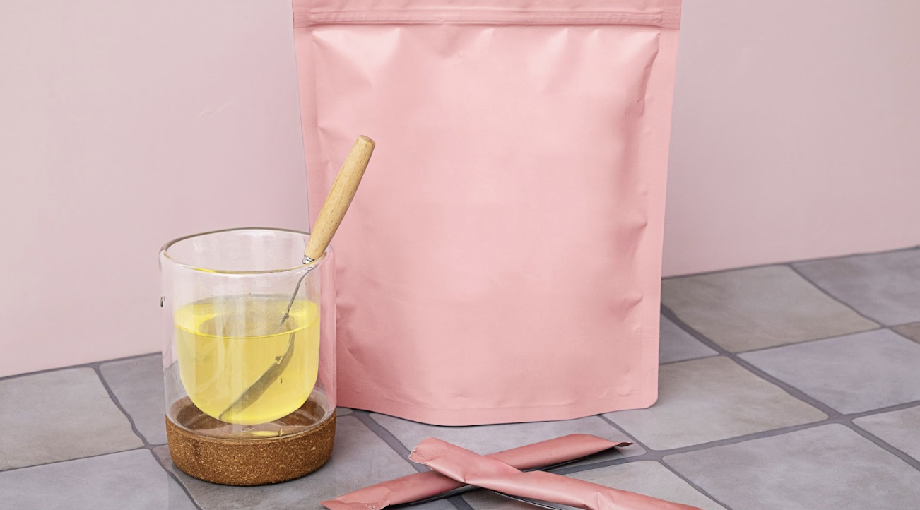 粉紅色大夾鏈袋包裝前，放著一杯裝有黃色液體的透明杯與攪拌匙，桌上還有兩條粉色隨身包，整體色調柔和，展現粉領族保養風格