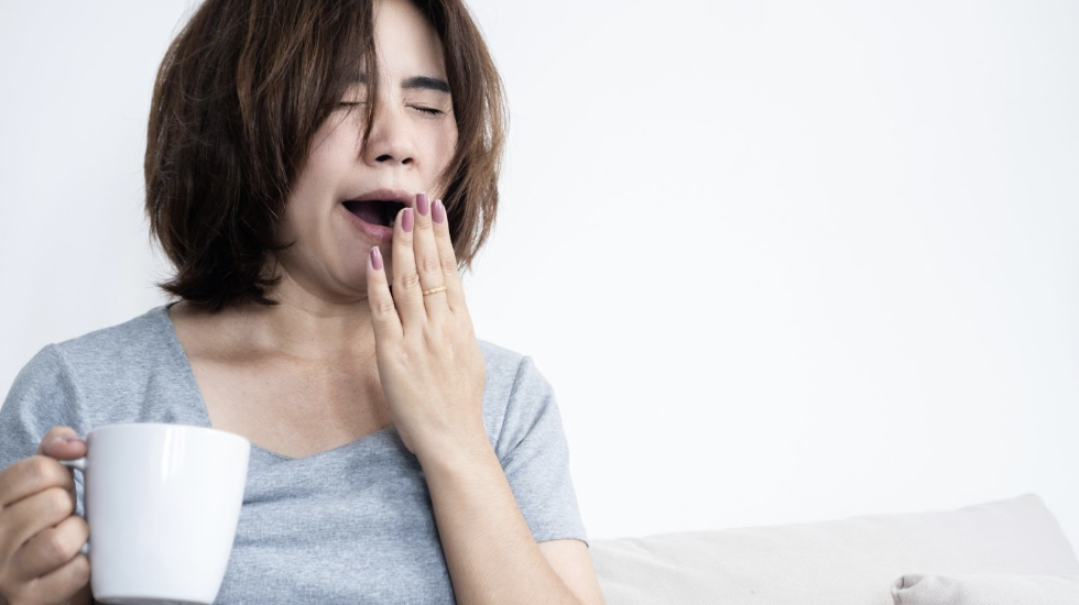 一位疲憊的女性手持白色馬克杯大聲打噴嚏或打哈欠，表現出體力不足與睏倦的狀態