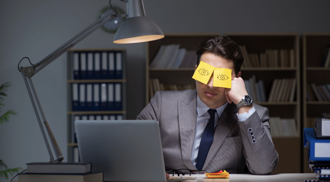男子在深夜辦公室加班，雙眼貼著畫有眼睛的黃色便利貼偽裝清醒。