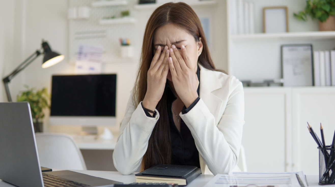 一名穿著白色西裝的職場女性在辦公桌前雙手按壓眉間，神情疲憊且雙眼酸澀。