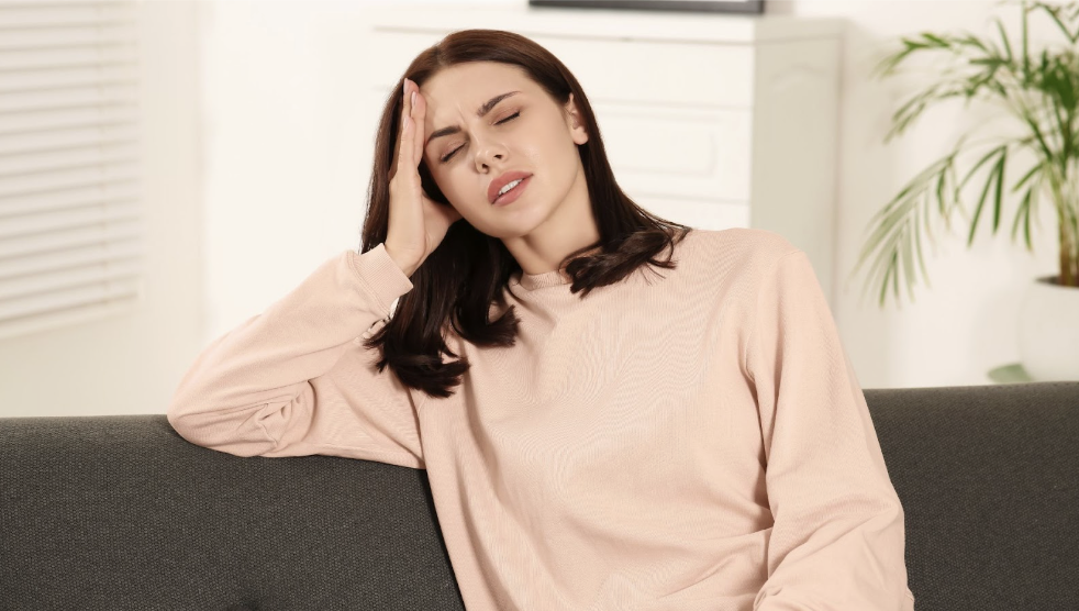 一名穿著粉色上衣的女性坐在沙發上,閉著眼睛並用手扶著額頭,表情顯得疲憊、頭痛或身體不適。
