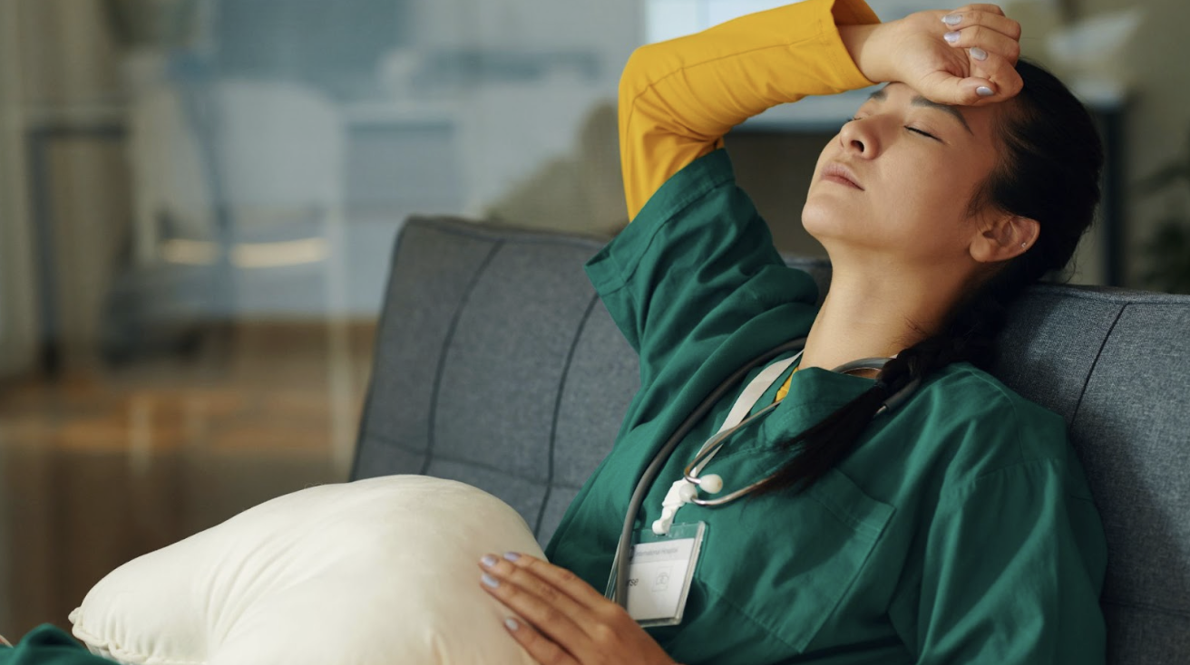 穿著綠色醫護服的女性醫護人員坐在沙發上休息,手扶額頭顯露疲憊與壓力狀態
