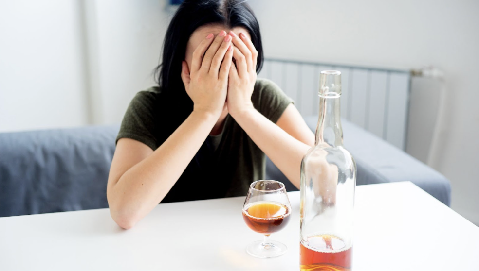 女性坐在桌前雙手遮臉顯得懊惱疲憊,桌上擺放酒瓶與半杯酒