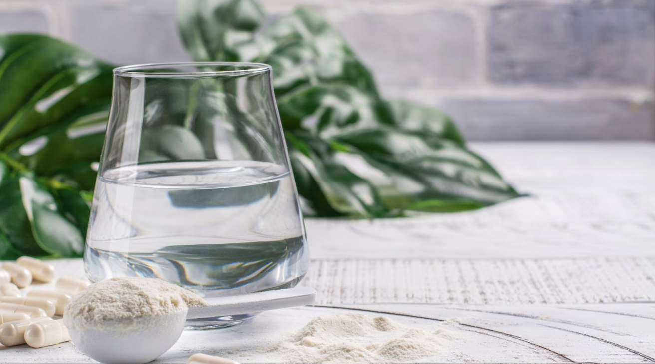 一杯純淨水旁放著一勺白色營養粉末與數顆透明膠囊，背景為綠色植物與灰白色磚牆，呈現健康生活的氛圍。