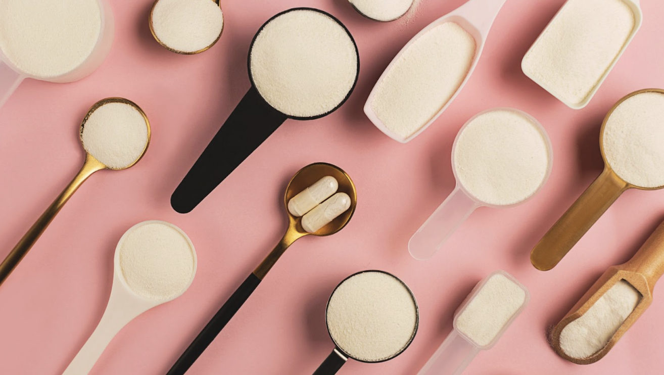 粉紅色背景上整齊排列著多種不同形狀與顏色的量勺，裝滿了白色粉末，其中一支金屬湯匙盛放著兩顆膠囊