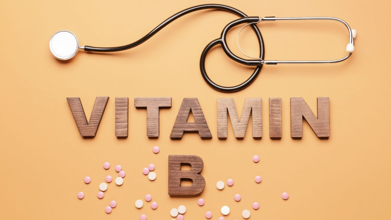 橘色背景上擺放著木製字母組成的「VITAMIN B」字樣,上方有一副專業醫療聽診器,周圍散布著粉色與白色的藥錠。