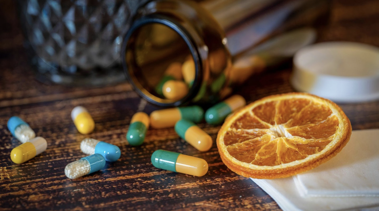 深色木紋桌面上散布著藍色與綠黃雙色的膠囊,旁邊有一片乾燥的橙片,背景是一個深色玻璃藥瓶,光影柔和。
