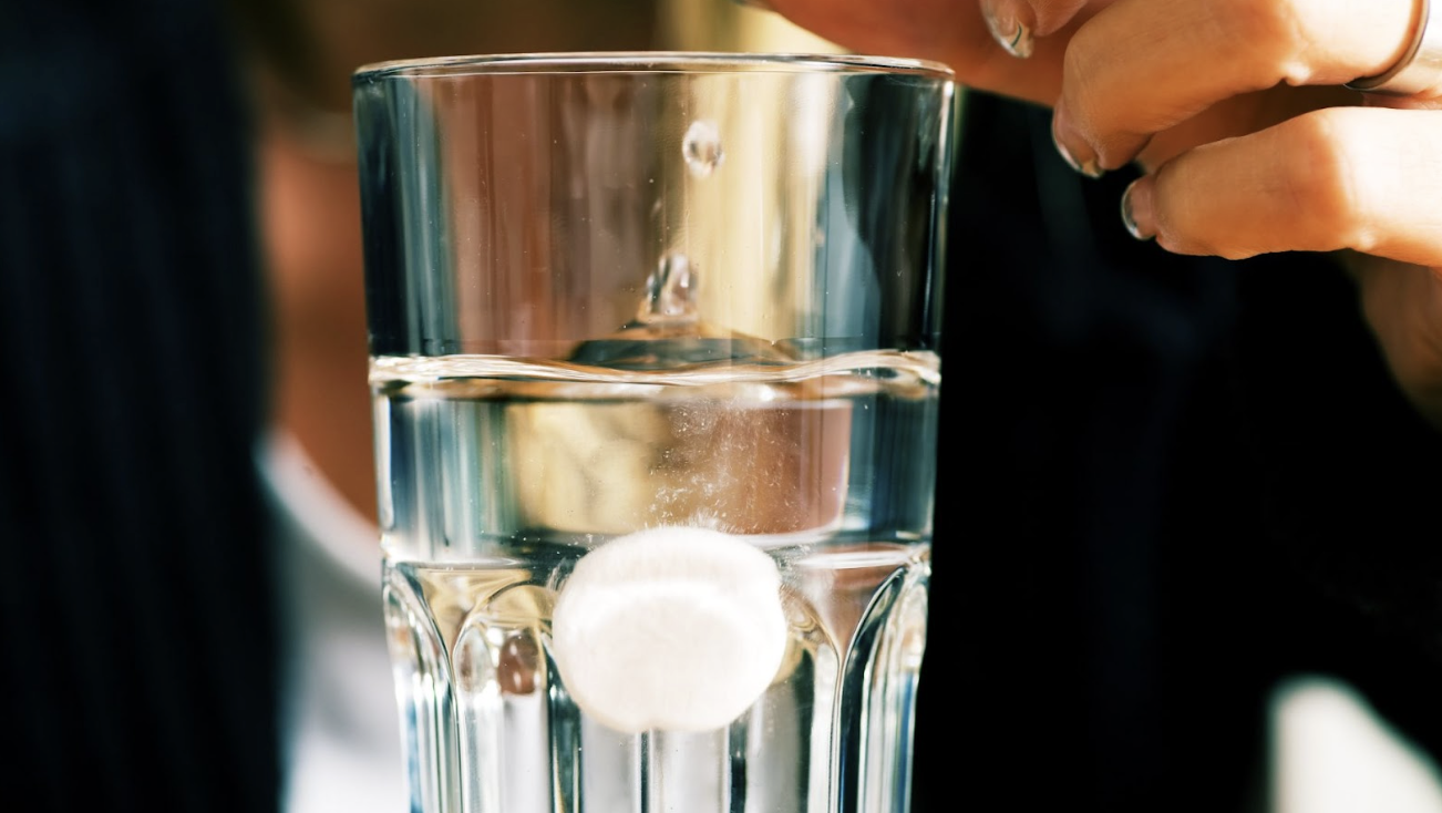 一顆白色的發泡錠正在透明水杯中迅速溶解,產生大量細小氣泡,水面有明顯的波動。