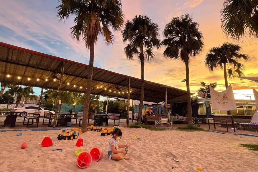 「請上車 5號港灣」景觀餐廳的兒童沙坑區，配有挖土機與小水桶玩具，背景為夕陽下的棕櫚樹