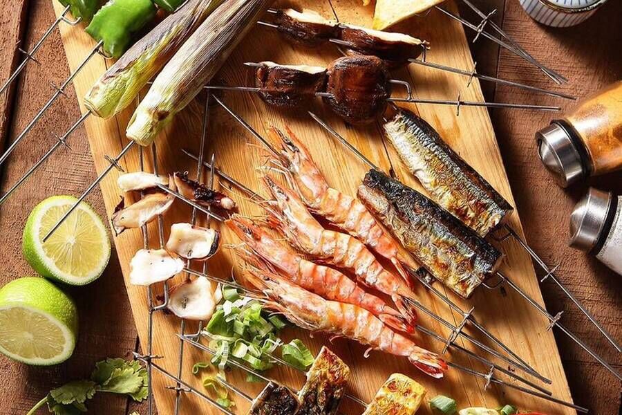 木質砧板上擺滿各式串燒海鮮與蔬菜，包括鮮蝦、秋刀魚、烤蔥段、雞翅與菇類，旁邊點綴檸檬與調味罐，呈現豐富誘人的燒烤拼盤。