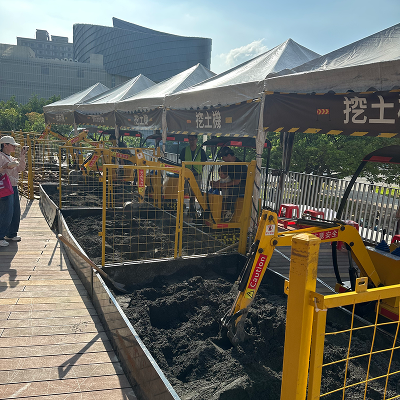 小型挖土機體驗區，孩子在沙坑中操作迷你挖土機挖沙，活動區設有帳篷遮陽與安全圍欄，戶外晴朗天氣下的親子互動遊戲場景。