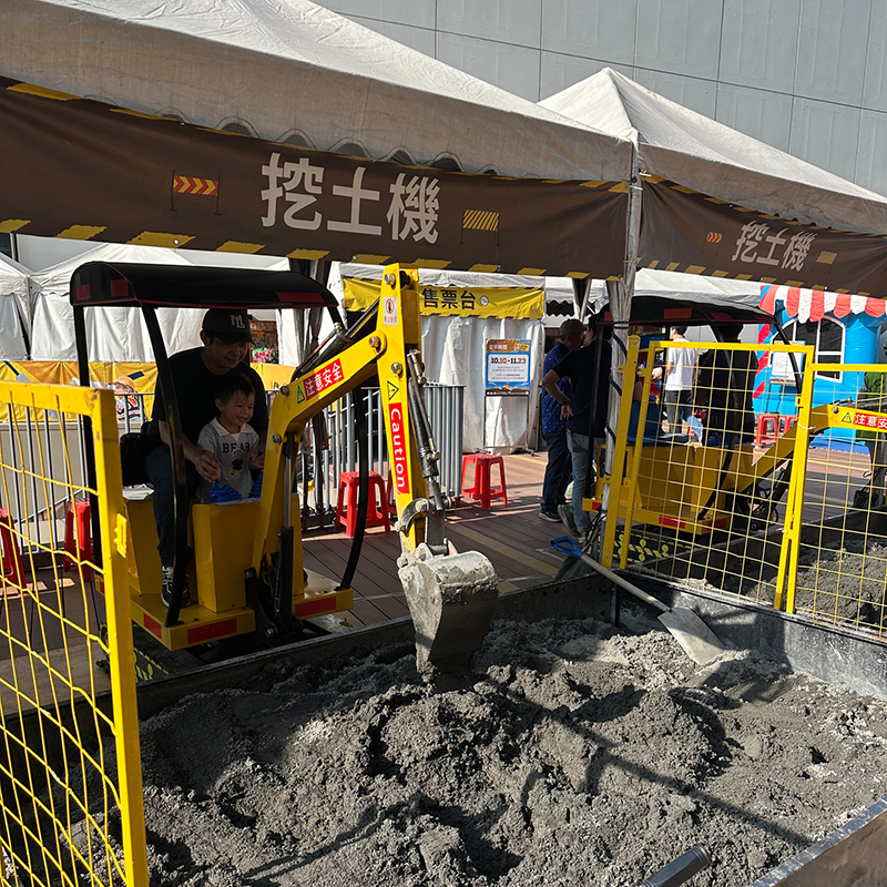 小朋友在家長陪同下體驗迷你挖土機挖沙活動，活動區設有帳篷與安全圍欄，上方標示「挖土機」體驗區，為戶外親子互動遊戲場景。