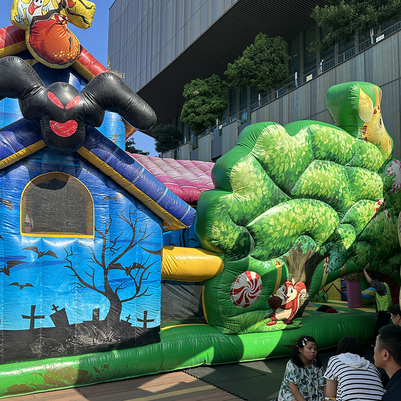 充氣式兒童遊樂設施，包含萬聖節鬼屋造型滑梯與森林主題充氣城堡，設置於戶外廣場，前方有家長與孩童休息。