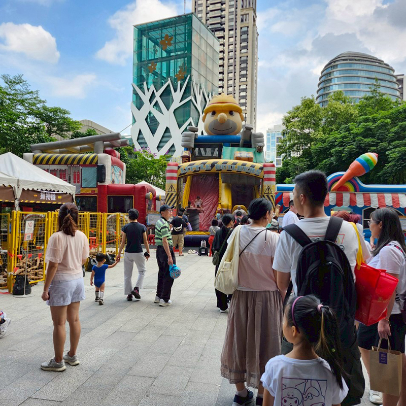 戶外親子活動現場，許多家庭排隊參加大型充氣工程主題遊戲設施，小朋友在建築工人造型充氣滑梯前遊玩，周圍設有挖土機體驗區與攤位，城市高樓與綠樹環繞的熱鬧場景。