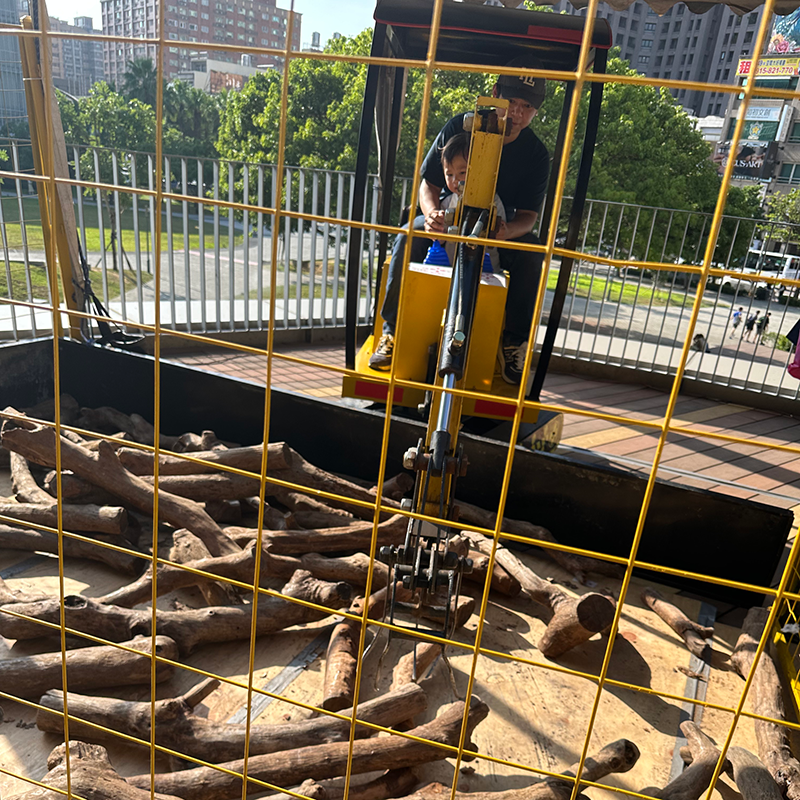 一名成人陪同幼童坐在小型挖土機操作座上，於戶外體驗區透過操縱桿挖取前方沙地中的木材，外圍設有黃色安全圍欄。