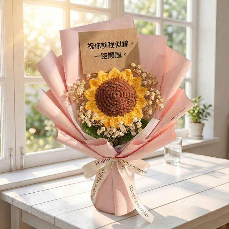 這張圖片的 ALT 替代文字建議如下：  明亮的白色木質桌面上，擺放著一束粉紅色包裝的手工編織向日葵花束。向日葵中心為深棕色，花瓣為金黃色，周圍點綴著白色滿天星乾花，並附有一張寫著「祝你前程似錦，一路順風」的棕色賀卡。背景為陽光灑落的白色窗戶與室內綠色小盆栽，營造出清新優雅的氛圍。