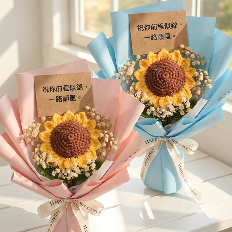 白色木桌上擺放著兩束精緻的編織向日葵花束，左邊為粉紅色包裝，右邊為淺藍色包裝。兩束花皆附有棕色小卡，印著「祝你前程似錦，一路順風」字樣，背景為明亮的白色窗台陽光。