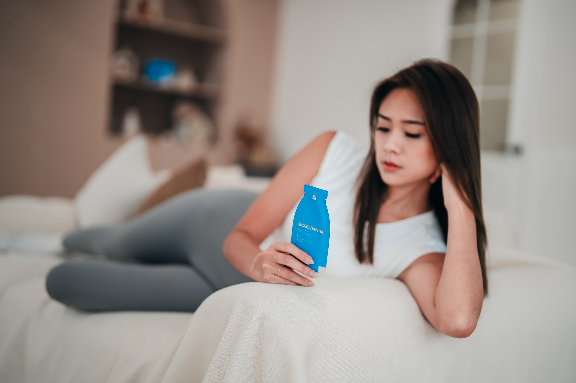 一位女性在居家沙發上側躺放鬆,手持藍色包裝的樂衛康玻潤凝口服玻尿酸飲品,呈現舒適自然的居家保養氛圍。