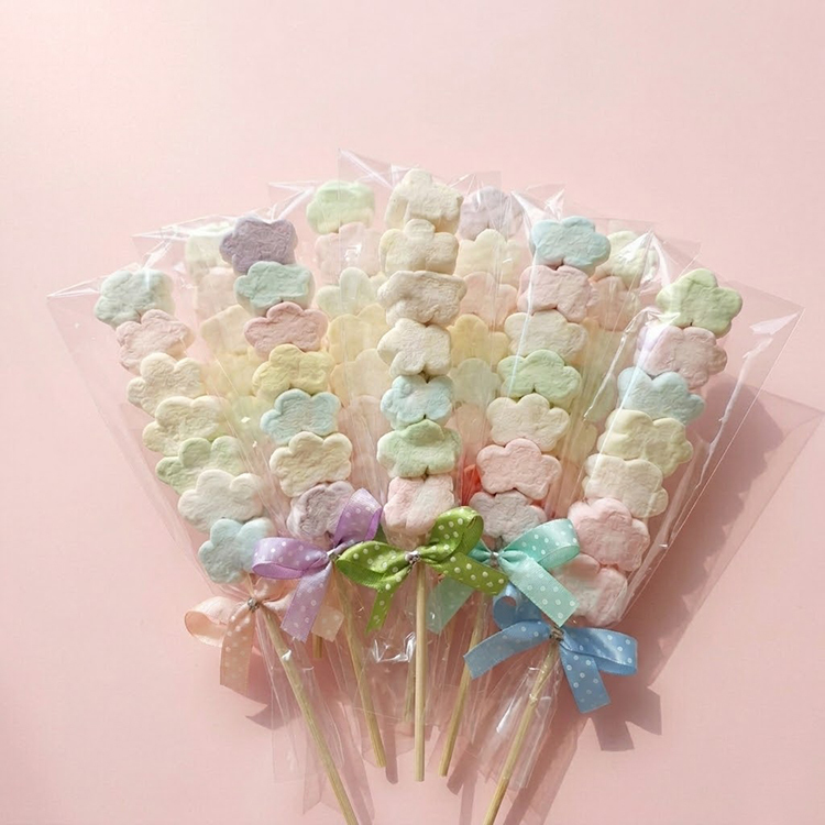 10支粉嫩色系花朵棉花糖串，搭配混色點點蝴蝶結，精緻包裝適合作為婚禮小物，背景為質感淡粉色 #FDEBED 桌面。