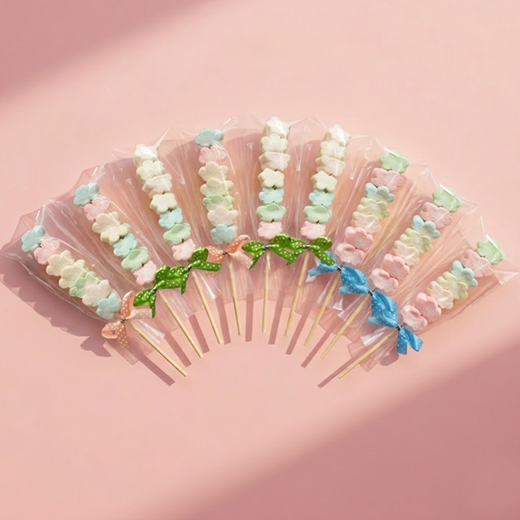 9支粉嫩色系花朵棉花糖串，搭配混色點點蝴蝶結，精緻包裝適合作為婚禮小物，背景為質感淡粉色 #FDEBED 桌面。