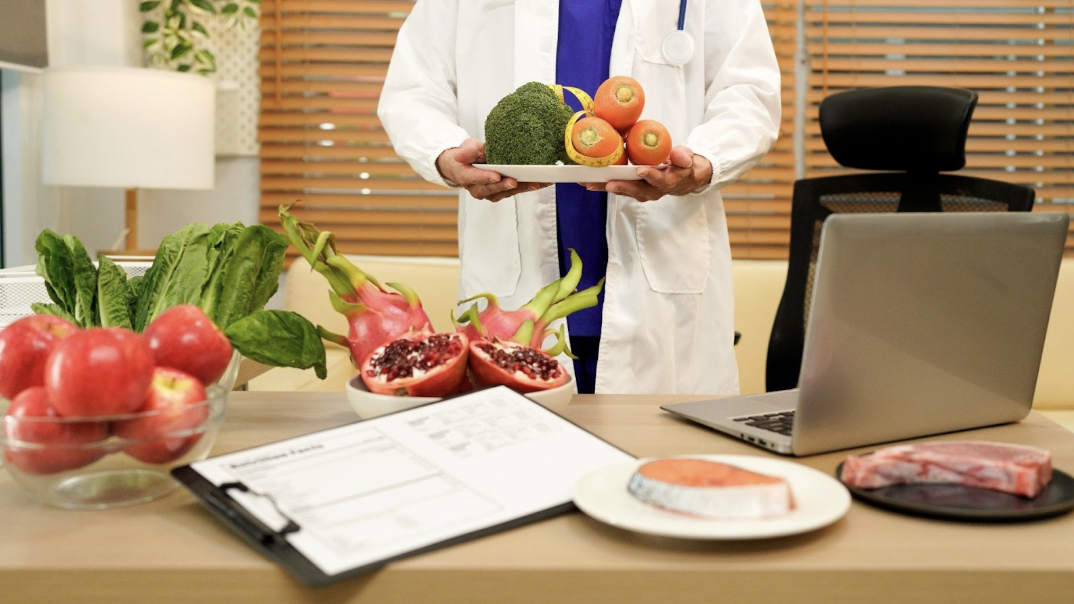 營養師身穿白袍展示裝滿花椰菜與紅蘿蔔的餐盤，桌上有火龍果、鮭魚與營養評估表。
