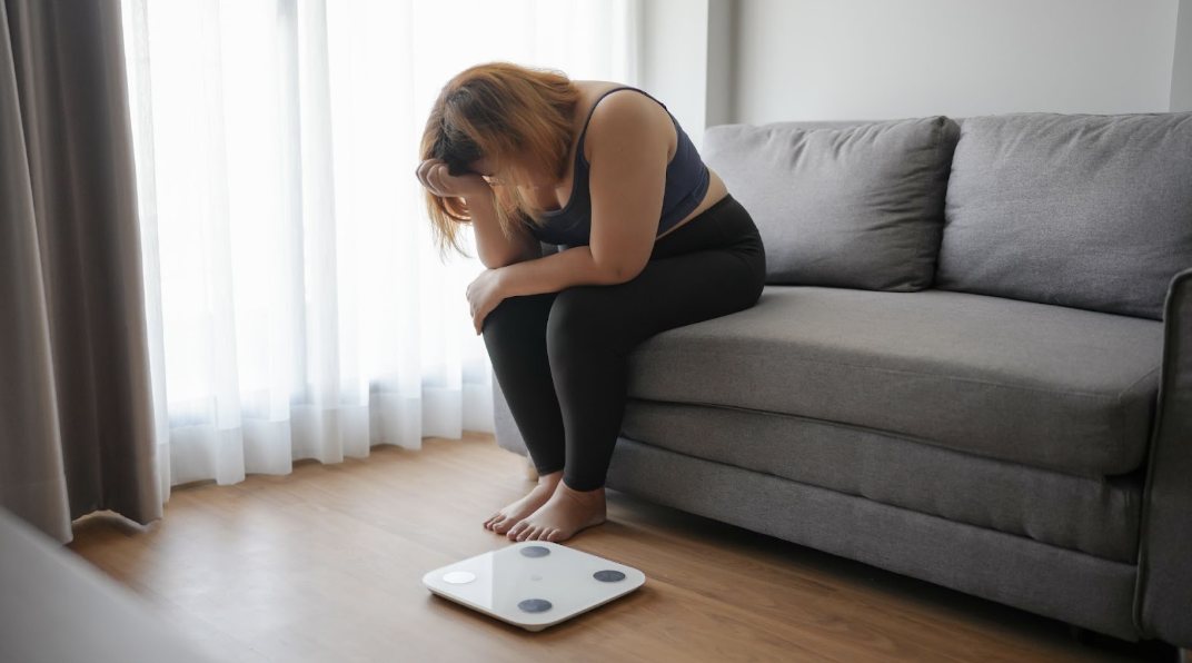 一位女性坐在沙發邊低頭苦惱,前方地板上放著體重計,呈現體重管理的壓力