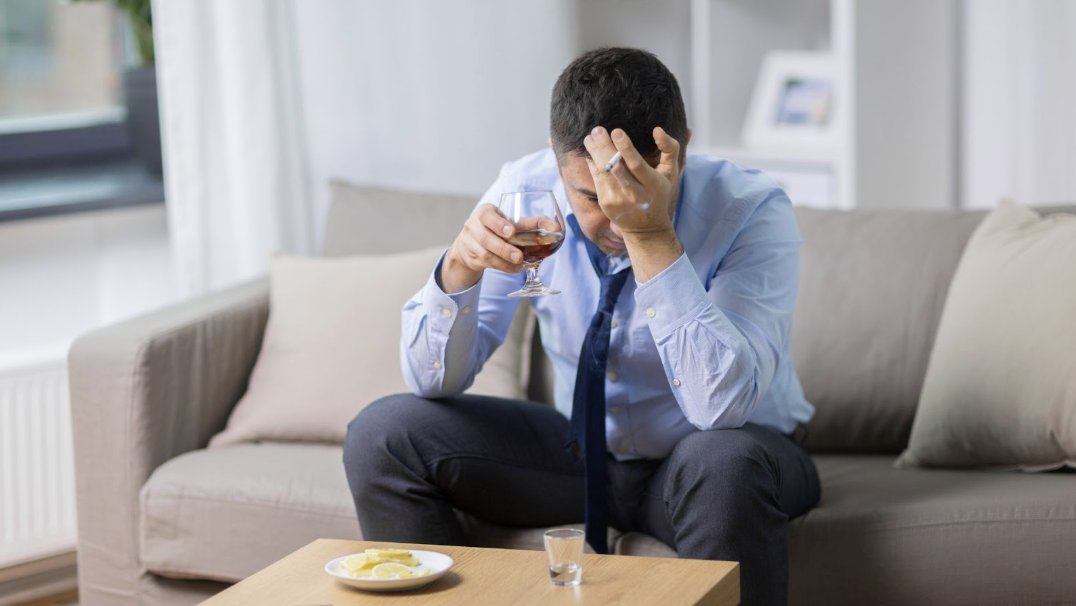 穿著襯衫與領帶的男子坐在沙發上，一手拿酒杯、一手扶額抽菸，顯現應酬疲憊與壓力