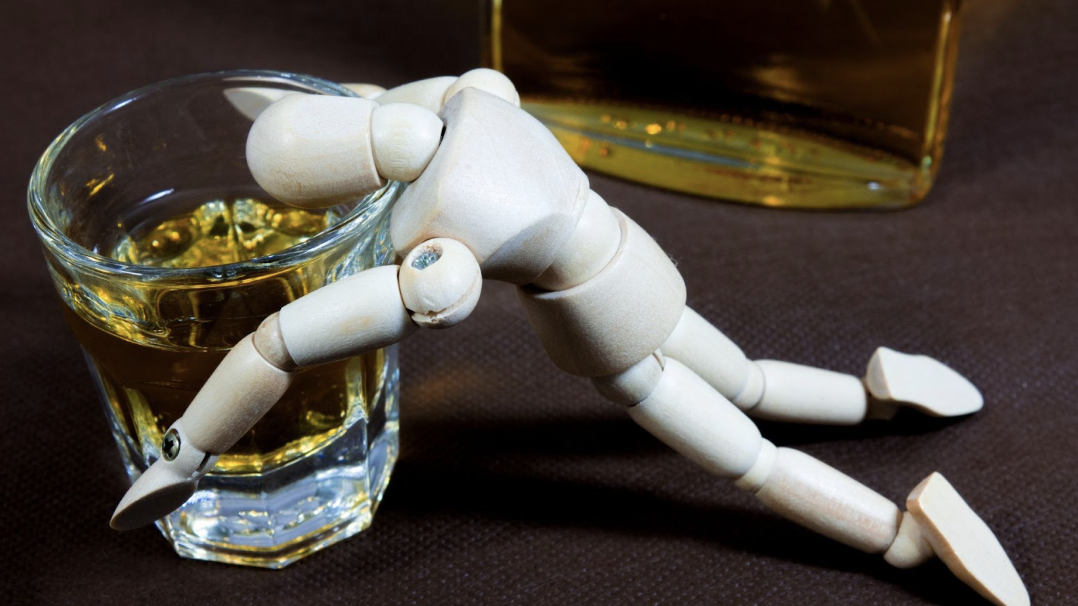 木頭關節人偶趴倒在裝有黃色液體的烈酒杯邊,旁邊有一瓶烈酒,暗喻過度飲酒後的疲態