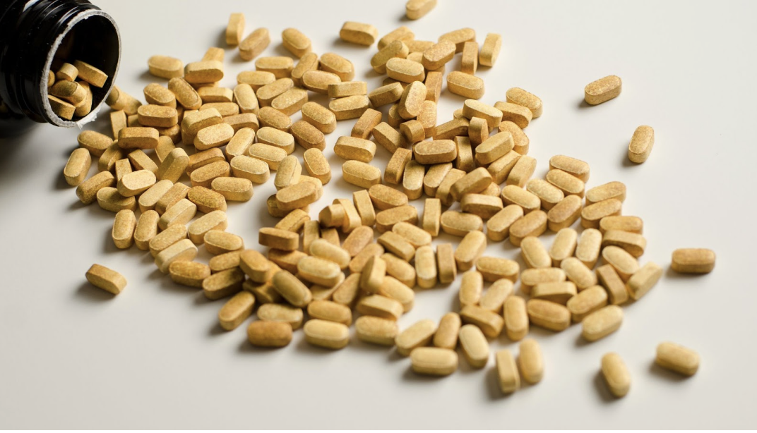 從深色藥瓶中灑出在白色平面上的大量淺橙色橢圓形營養補充品錠劑。