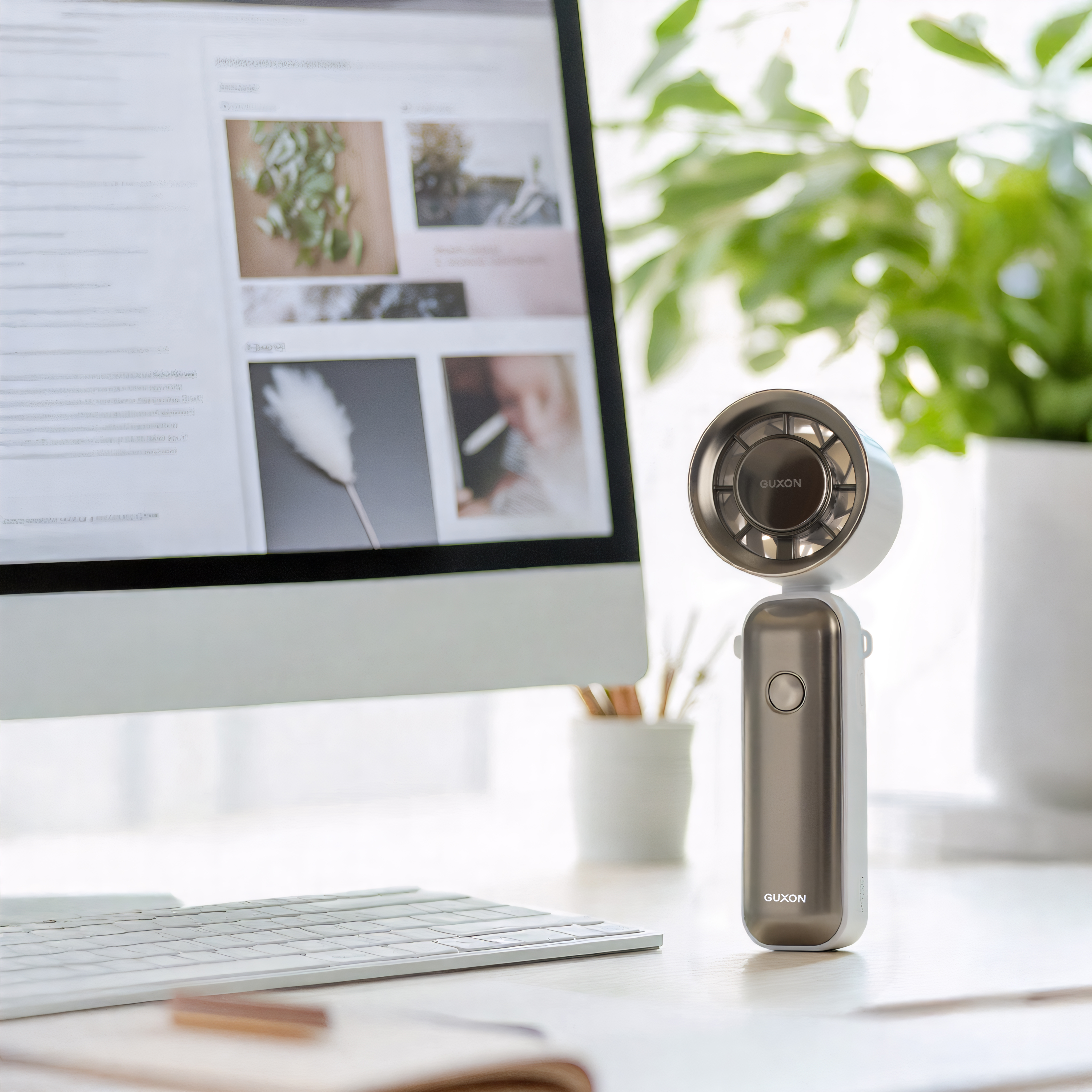Silver-grey GUXON handheld fan beside a laptop and keyboard by a window