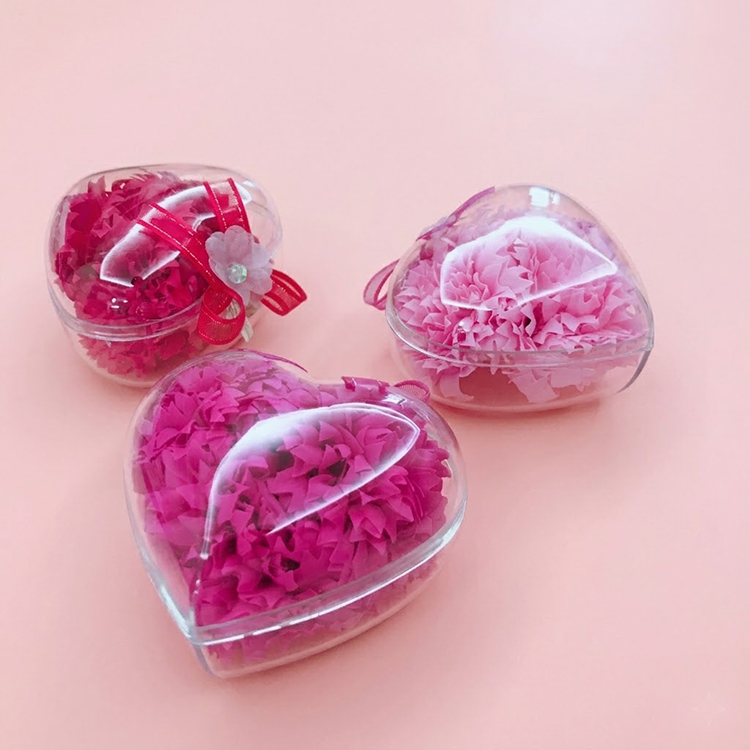 一張俯拍照片，展示了三個心形的透明壓克力盒，放置在柔和的淺粉色背景上。左上角的盒子裡裝著鮮紅色的香皂花，並裝飾著紅色的絲帶和一朵白色小花。右邊的盒子裡裝著淺粉色的香皂花，而底部的盒子裡則裝著亮粉色的香皂花。