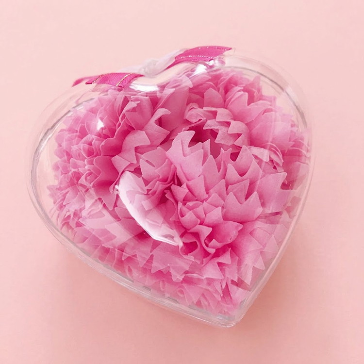 一個心形的透明塑膠盒，裡面裝滿了粉紅色的紙製康乃馨，花瓣層次分明且帶有鋸齒狀邊緣。盒子的頂部裝飾著一條帶有金邊的亮粉色絲帶。整件物品放置在純淨且柔和的淺粉色（色碼 #FDEBED）背景中央。